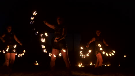 Three-women-with-burning-hoops-dance-with-fiery-torches-in-leather-clothes-in-a-dark-hangar-demonstrating-a-circus-fire-show-in-slow-motion